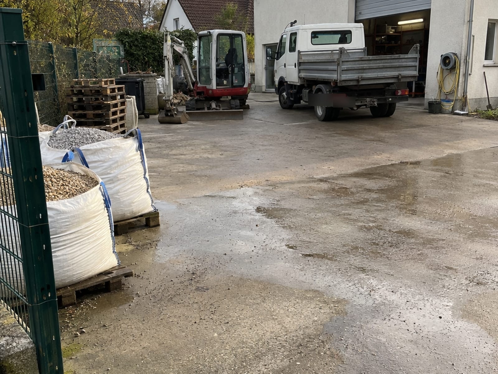 Betriebshof mit Minibagger, Transporter und Materialsäcken vor der Werkstatt