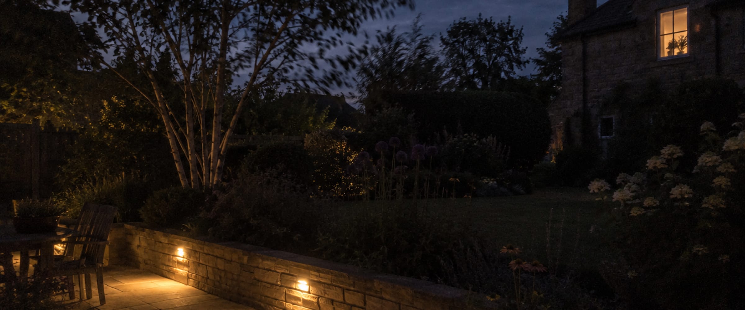 Garten in der Dämmerung mit beleuchteter Sitzmauer und warmem Lichtschein aus dem Haus
